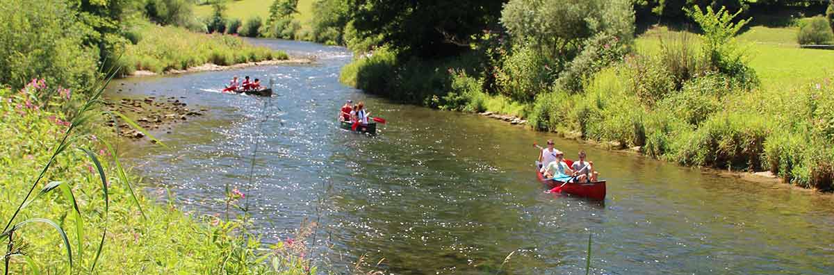 3 vollbesetzte Kanus auf dem oberen Neckar, vom rechten Ufer aus fotografiert; der Fluss schlängelt sich von einer Biegung im Hintergrund über eine zweite nach vorn