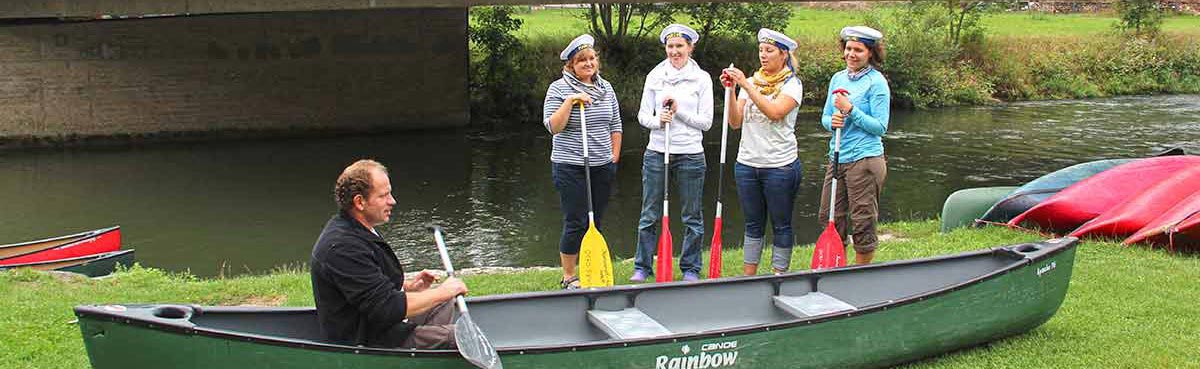 Einweisung Einweisung. Kanu am Ufer, Einweiser sitzt mit Paddel im Boot, hinter dem Kanu 2 Frauen mit Paddeel am rechten Bildrand; dritte Frau am Rand halb verdeckt. Dahinter Neckar und Brückenpfeiler.