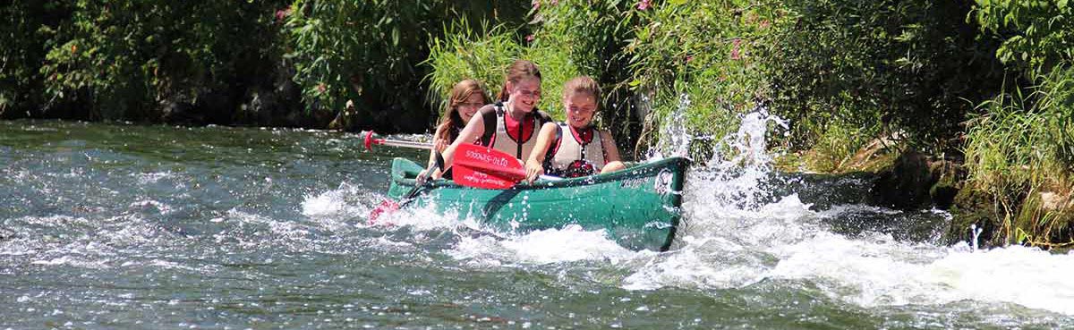Flotte Fahrt Flotte Fahrt. Drei Mädels fahren durch Stromschnelle in einem grünen Kanu. Blick von schräg vorne auf Boot am rechten Bildrand