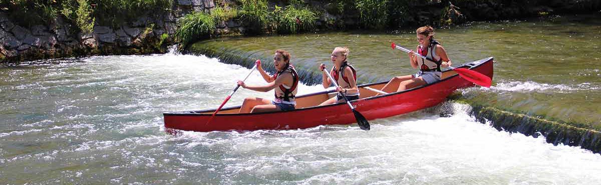 Staustufe-2 Rotes Kanu mit 2 paddelnden Frauen hat die Staustufe fast durchfahren; fahren vom rechten Bildrand ins Bild; gischtiges Wasser, dahinter Ufer mit Steinwand und Gebüsch